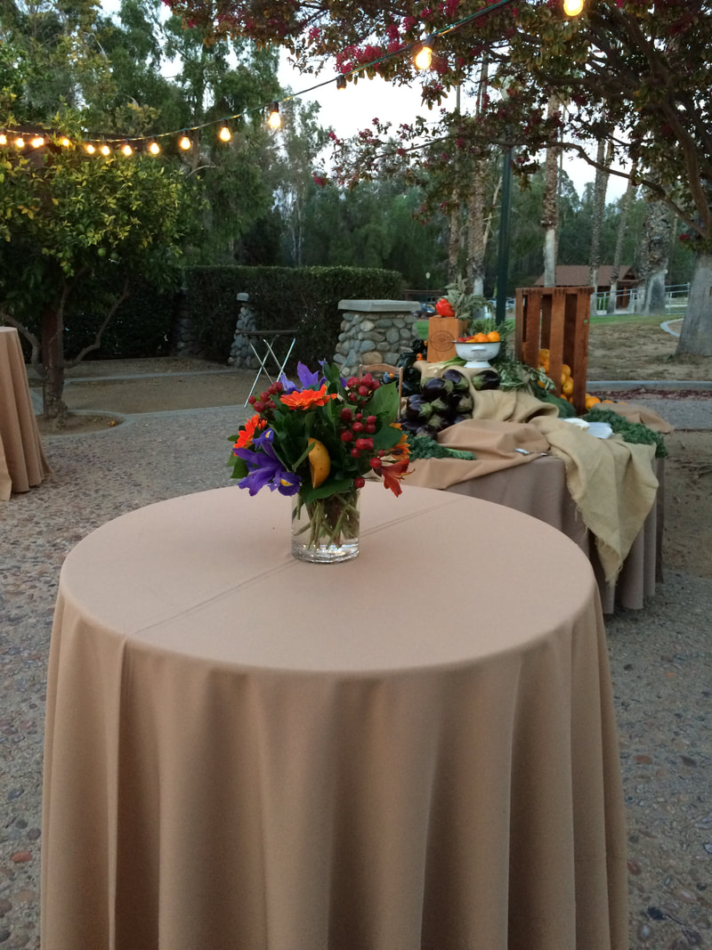 Picture of cocktail table with citrus-themed centerpiece, and a buffet station in the background, with trees and market lights at the courtyard of the Sunkist Room at the California Citrus State HIstoric Park in Riverside