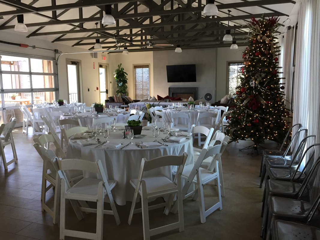 Picture of all-white, round guest tables and chairs inside a community center with a decorated Christmas tree in the room