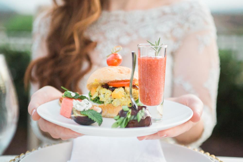 Picture of a bride holding a plate filled with appetizers: stuffed fig, marinated mushrooms, gazpacho shot glass, curried chicken slider, and watermelon bite